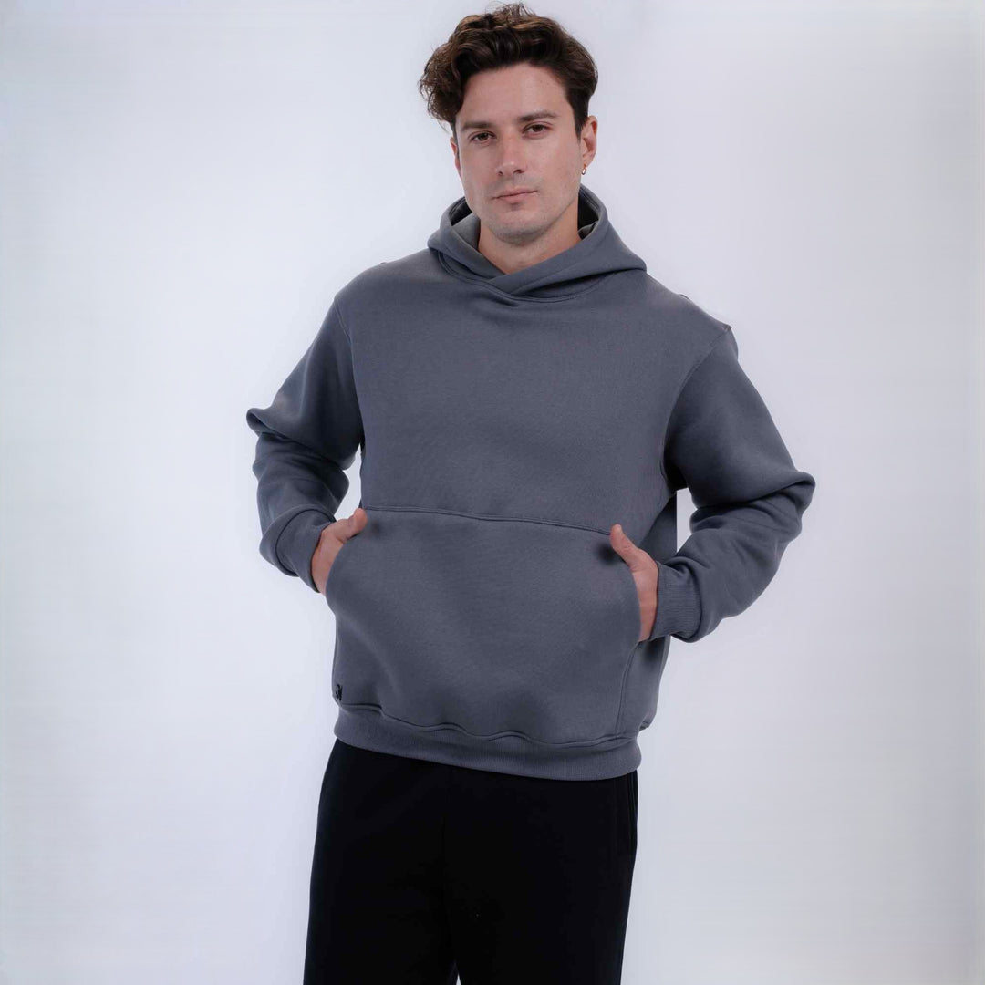 A man wearing a dark gray pullover hoodie with a front kangaroo pocket and black pants, standing against a light gray studio background.
#color_gray