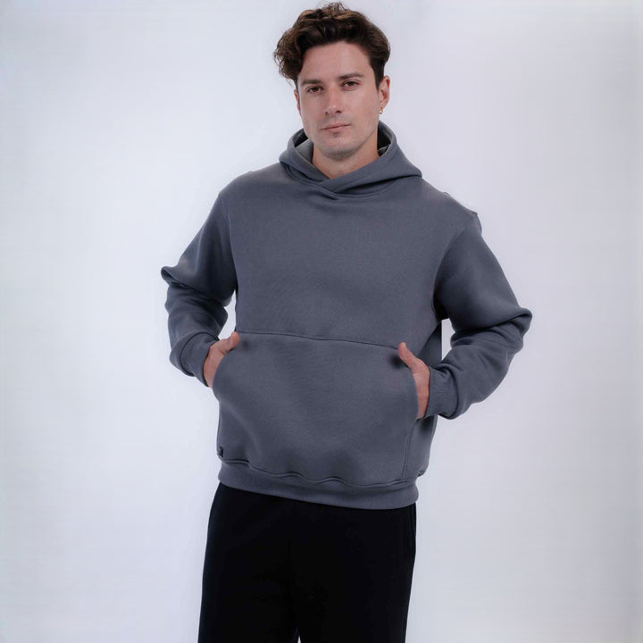 A man wearing a dark gray pullover hoodie with a front kangaroo pocket and black pants, standing against a light gray studio background.
#color_gray