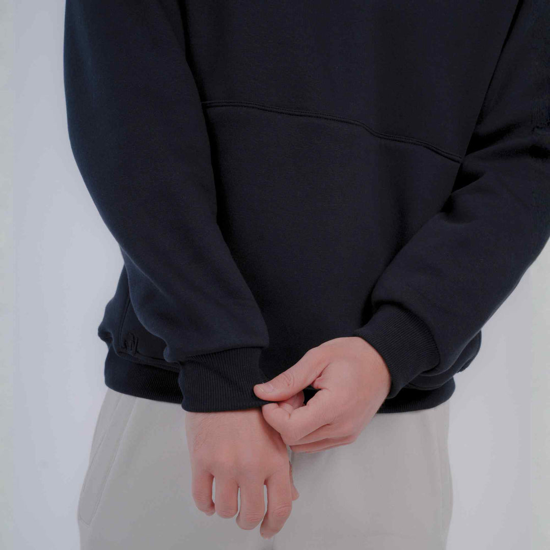 A close-up of a person wearing a black pullover sweatshirt and light beige pants, with the hands adjusting the cuff of the sweatshirt.
#color_black