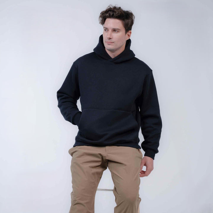 A man wearing a black pullover hoodie and tan pants, standing against a plain light gray studio background.
#color_black