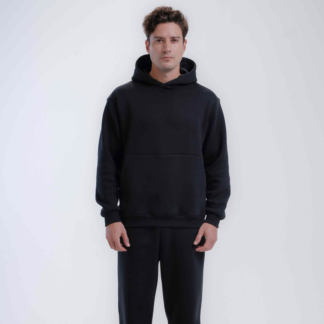 A man wearing a black pullover hoodie with a front kangaroo pocket and matching black sweatpants, standing against a light gray studio background.
#color_black
