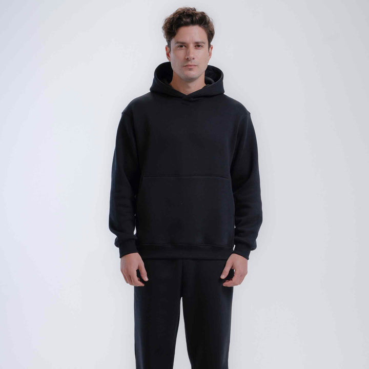 A man wearing a black pullover hoodie with a front kangaroo pocket and matching black sweatpants, standing against a light gray studio background.
#color_black