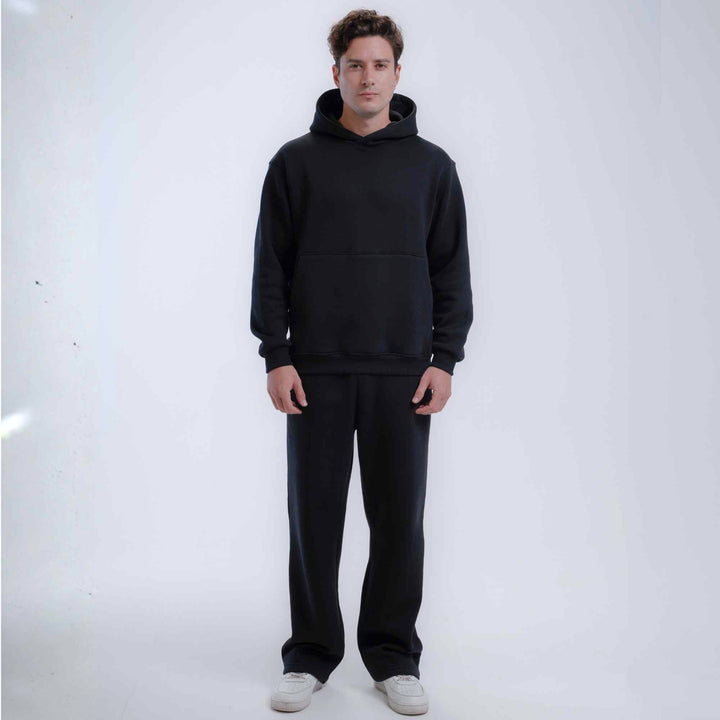 A man modeling a black pullover hoodie and matching black sweatpants against a plain white studio 
background.
#color_black