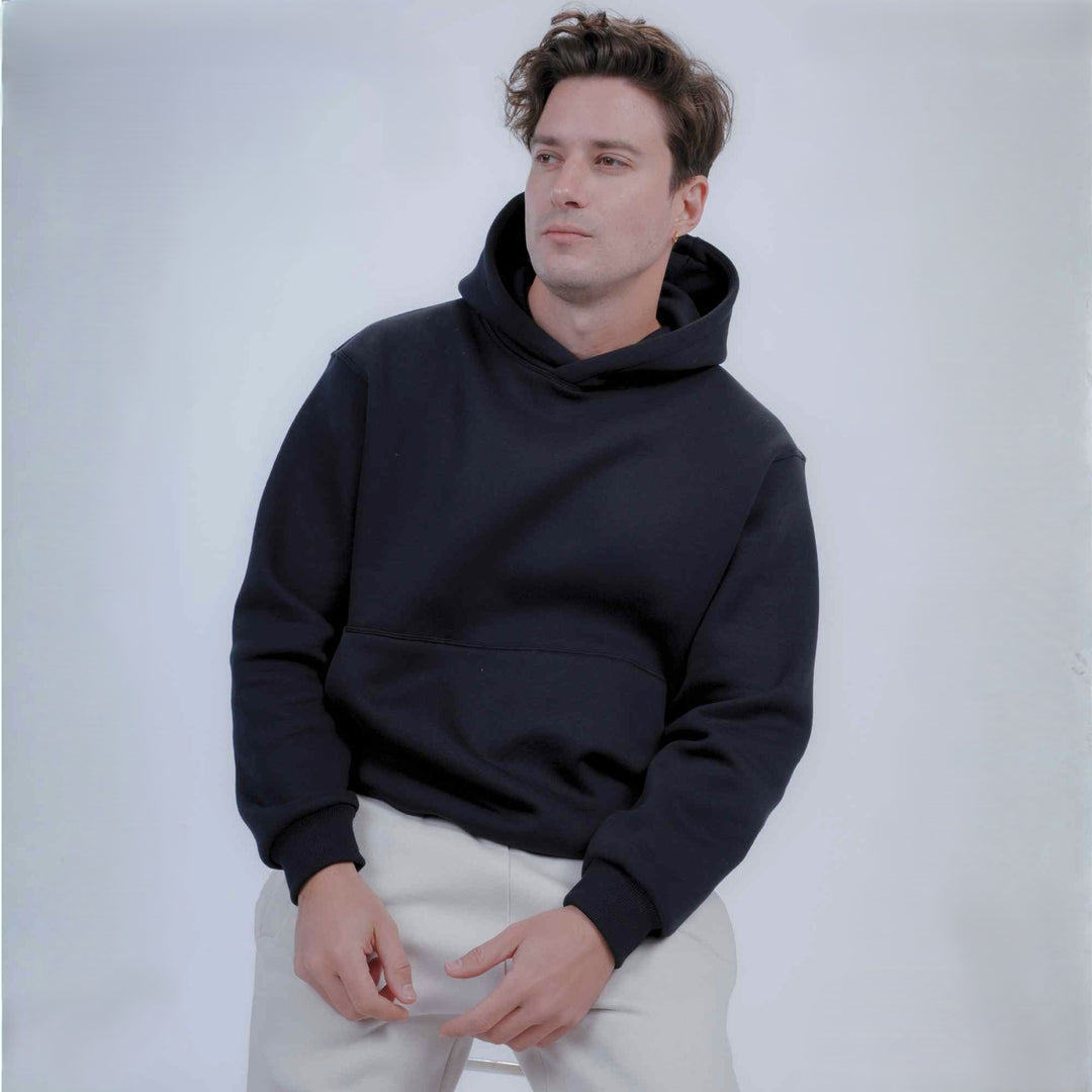 A man wearing black pullover hoodie with a front kangaroo pocket and light beige pants, standing against a light gray studio background.
#color_black