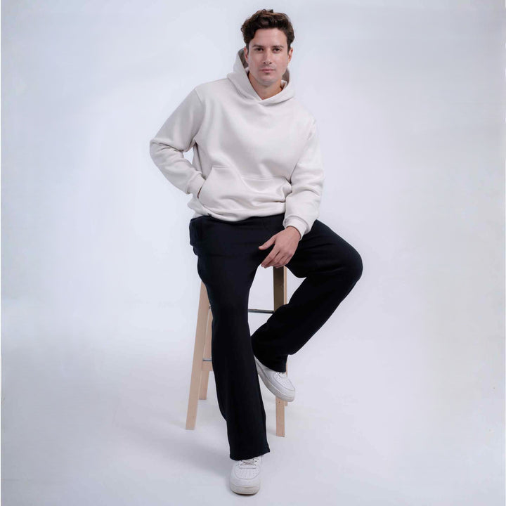 A man wearing a light cream-colored hoodie and black pants sits on a wooden stool against a light gray studio background.
#color_cloud-dancer
