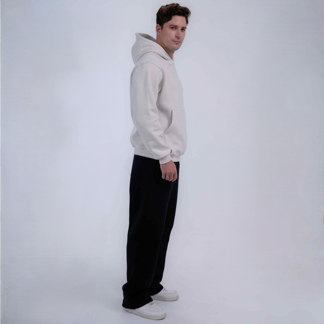 A man wearing a light beige/cream pullover hoodie, black wide-leg pants, and white sneakers, standing in profile against a plain light gray studio background.
#color_cloud-dancer