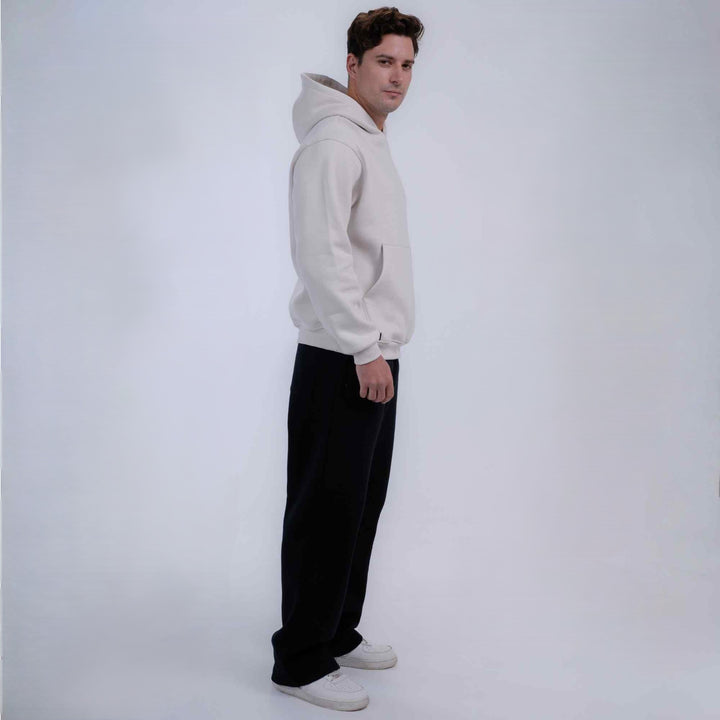 A man wearing a light beige/cream pullover hoodie, black wide-leg pants, and white sneakers, standing in profile against a plain light gray studio background.
#color_cloud-dancer