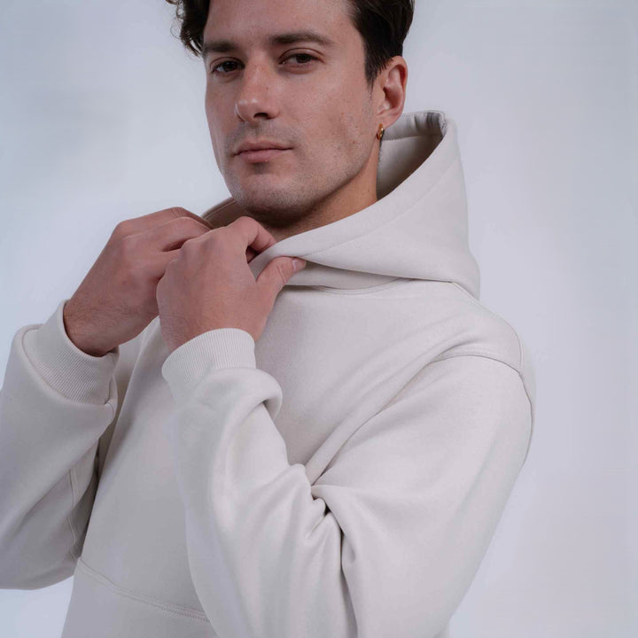 A man wearing a light beige or off-white hoodie, adjusting the collar with both hands against a plain light gray studio background.
#color_cloud-dancer