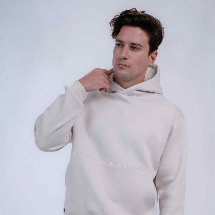 A man wearing a light beige oversized pullover hoodie, posing with one hand adjusting the hood against a plain light gray background.
#color_cloud-dancer