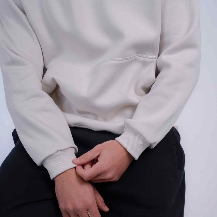 A close-up of a person wearing a light cream or off-white pullover hoodie with black pants, sitting against a plain light background.
#color_cloud-dancer