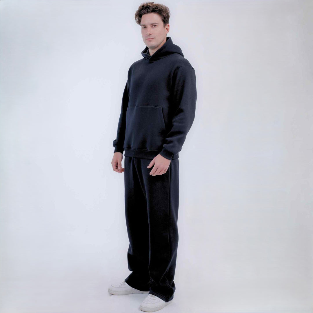 A man modeling a matching black hoodie and sweatpants set with white sneakers, standing against a plain light gray studio background.
#color_black