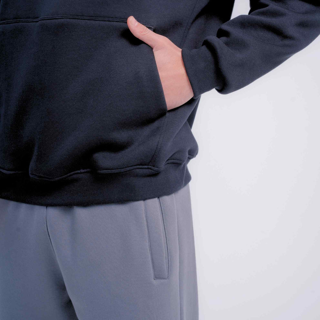 Close-up of a person wearing a dark navy pullover hoodie with a front pocket, with one hand in the pocket, paired with light gray pants against a plain light background.
#color_gray