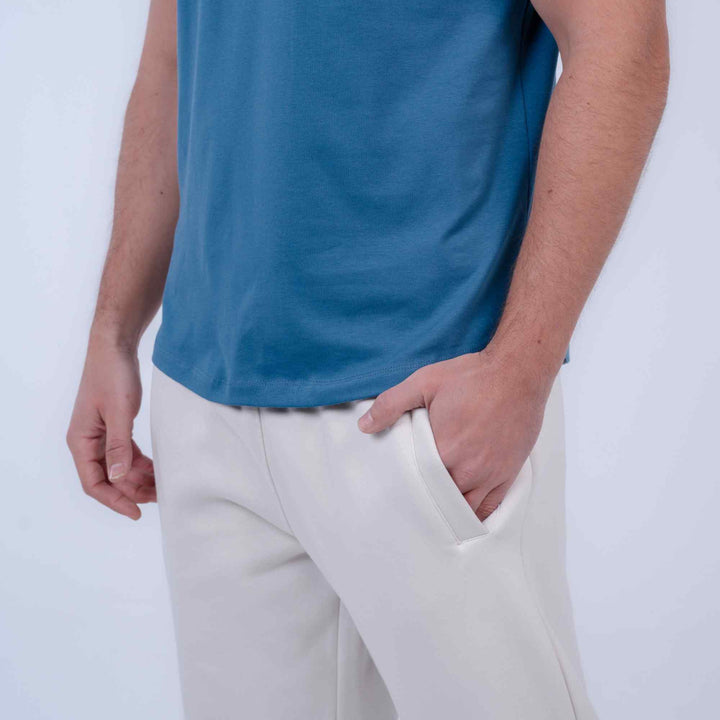A close-up of a person wearing a blue short-sleeve t-shirt and off-white trousers, with one hand in a pocket, against a plain light background.
#color_cloud-dancer