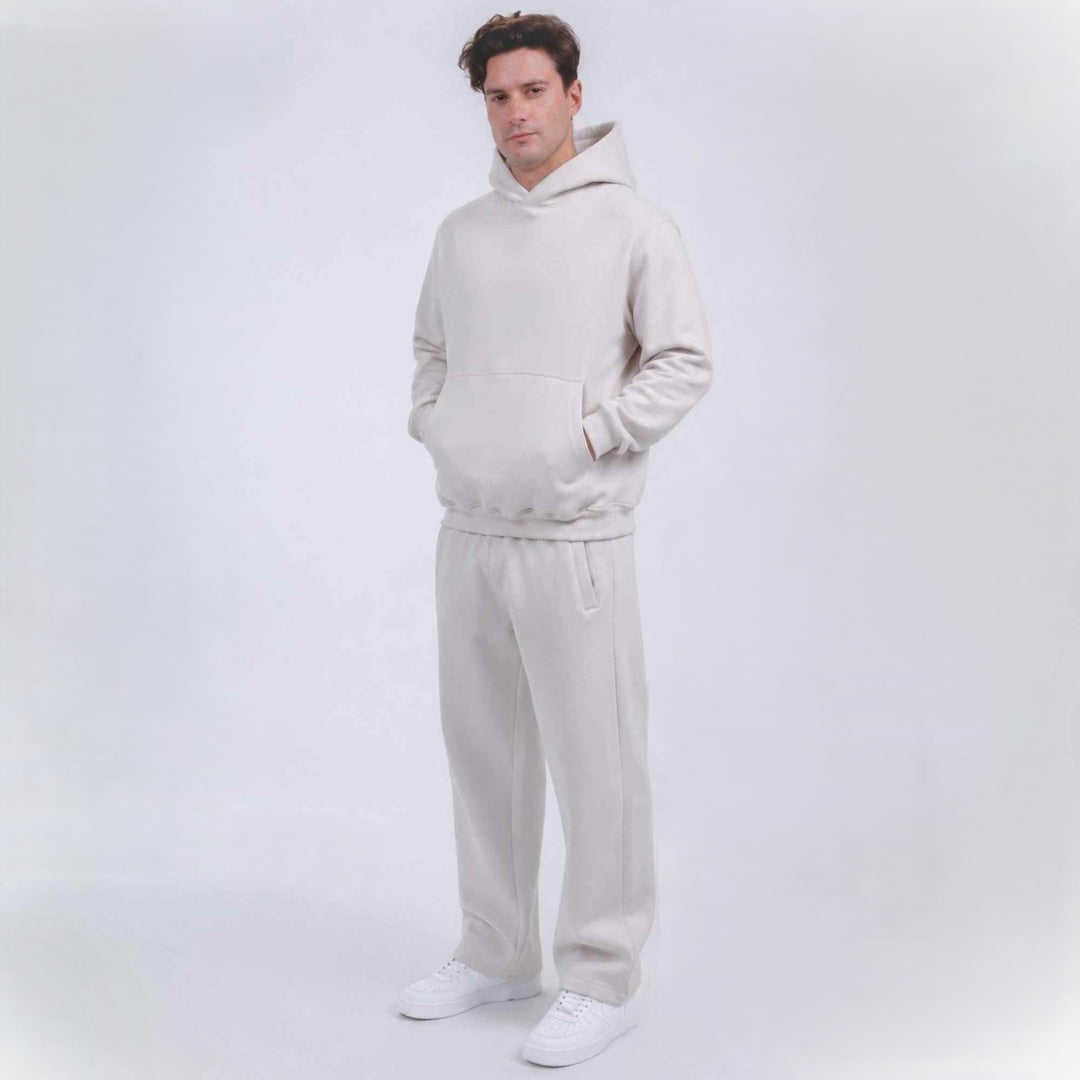 A man modeling a matching light beige hoodie and sweatpants set with white sneakers, standing against a seamless light gray studio background.
#color_cloud-dancer