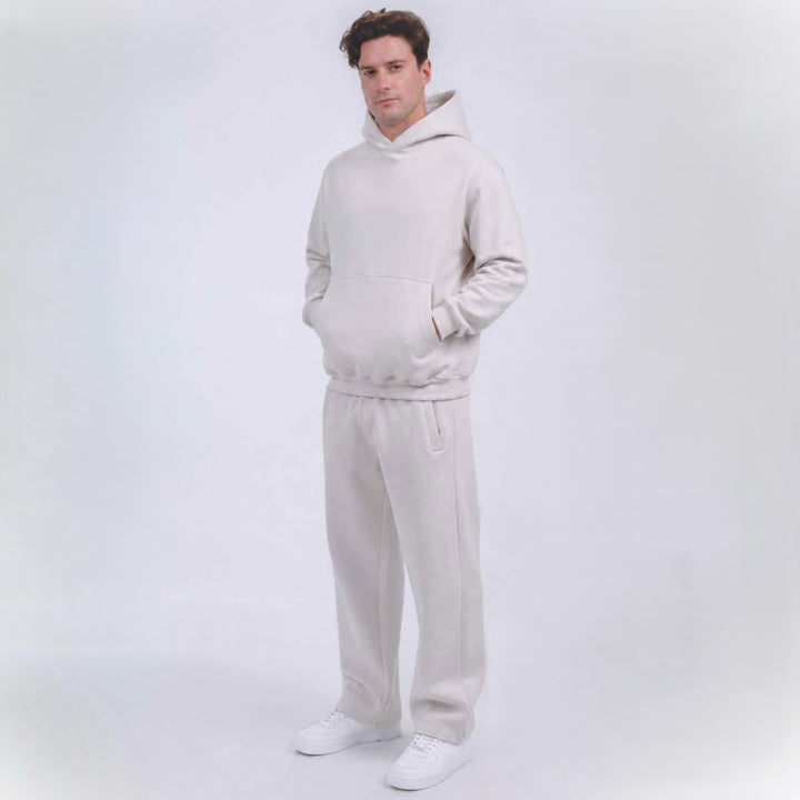 A man modeling a matching light beige hoodie and sweatpants set with white sneakers, standing against a seamless light gray studio background.
#color_cloud-dancer