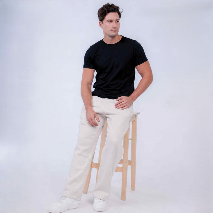 A man wearing a black short-sleeve crewneck t-shirt, off-white wide-leg trousers, and white sneakers, standing next to a light wood stool against a light gray studio background.
#color_cloud-dancer