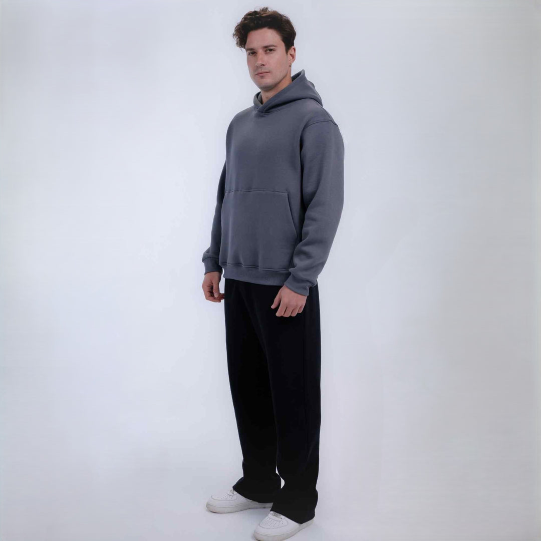 A man modeling a dark gray pullover hoodie with a front kangaroo pocket, paired with black sweatpants and white sneakers, standing against a plain white studio background.
#color_Grey