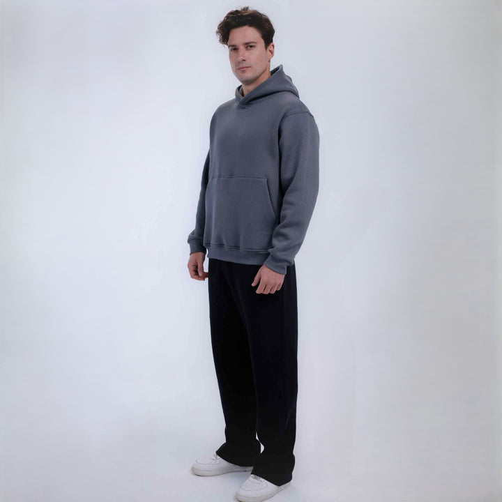 A man modeling a dark gray pullover hoodie with a front kangaroo pocket, paired with black sweatpants and white sneakers, standing against a plain white studio background.
#color_Grey