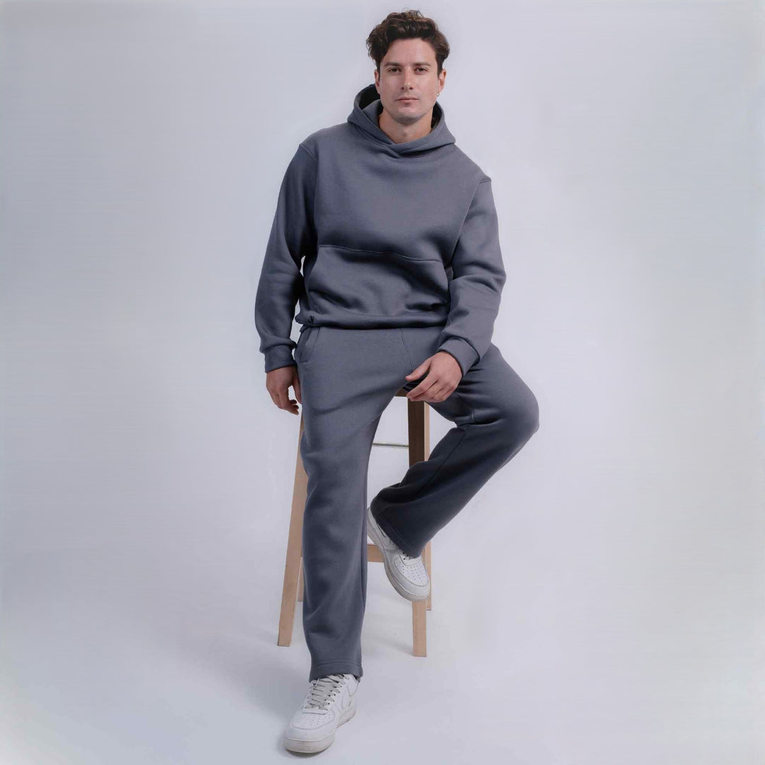 A man wearing a matching gray two-piece lounge set consisting of a pullover hoodie and sweatpants, sitting on a wooden stool against a light gray studio background.
#color_gray