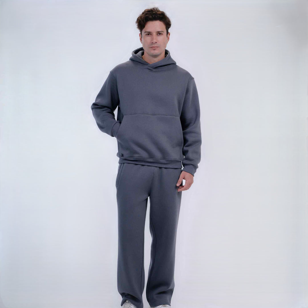 A man modeling a matching dark gray two-piece loungewear set, consisting of a pullover hoodie with a kangaroo pocket and matching sweatpants, against a plain light gray studio background.
#color_gray